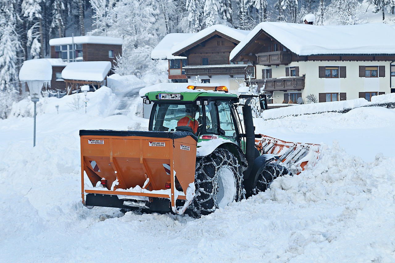 Traktor odśnieża zasypaną śniegiem drogę w g&oacute;rskiej scenerii. W tle ośnieżone domy i drzewa.