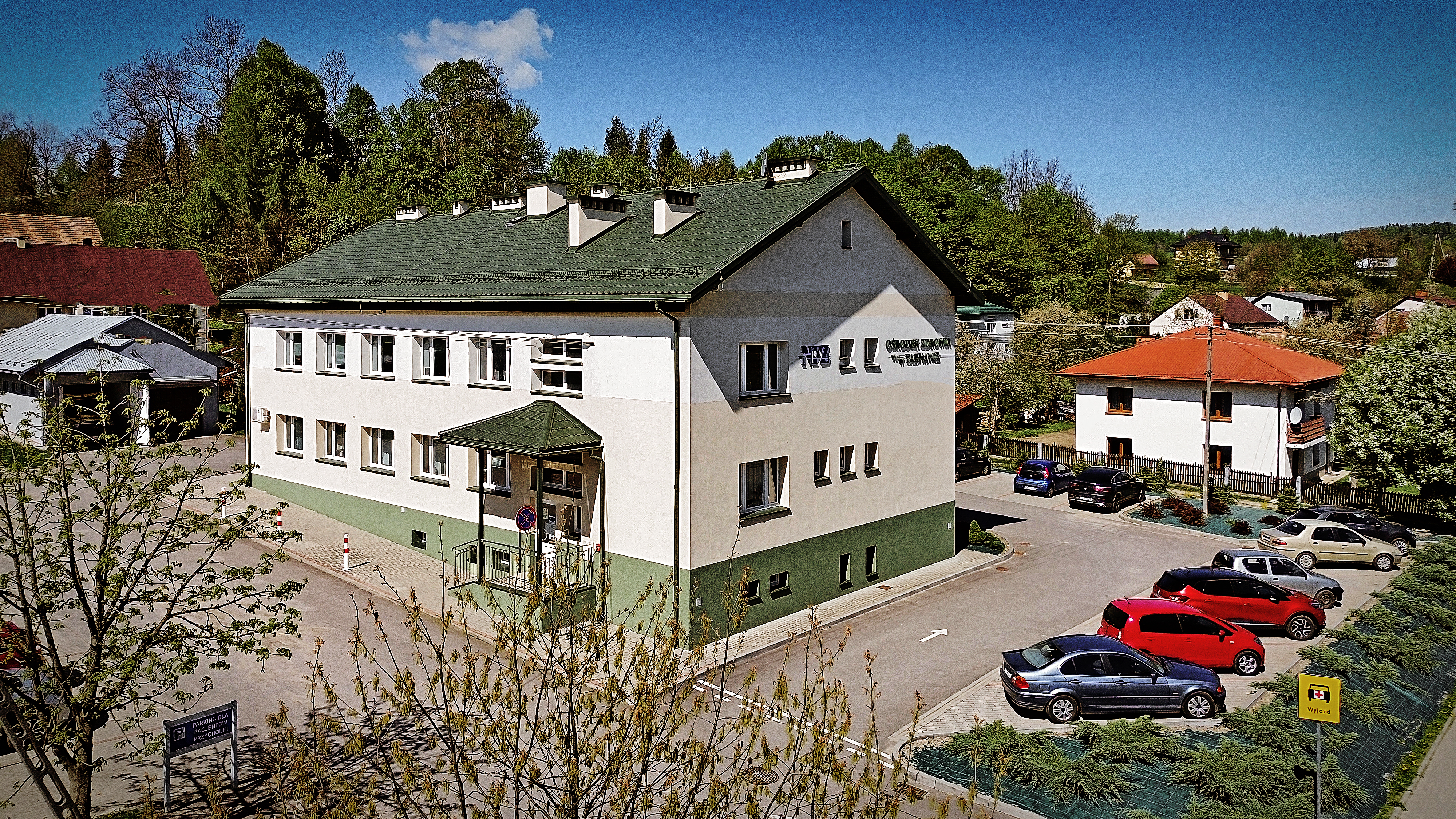 Zdjęcie budynku z zielonym dachem, prawdopodobnie ośrodka zdrowia, z parkingiem pełnym samochodów. Widoczne drzewa i inne domy w tle. Słoneczny dzień.