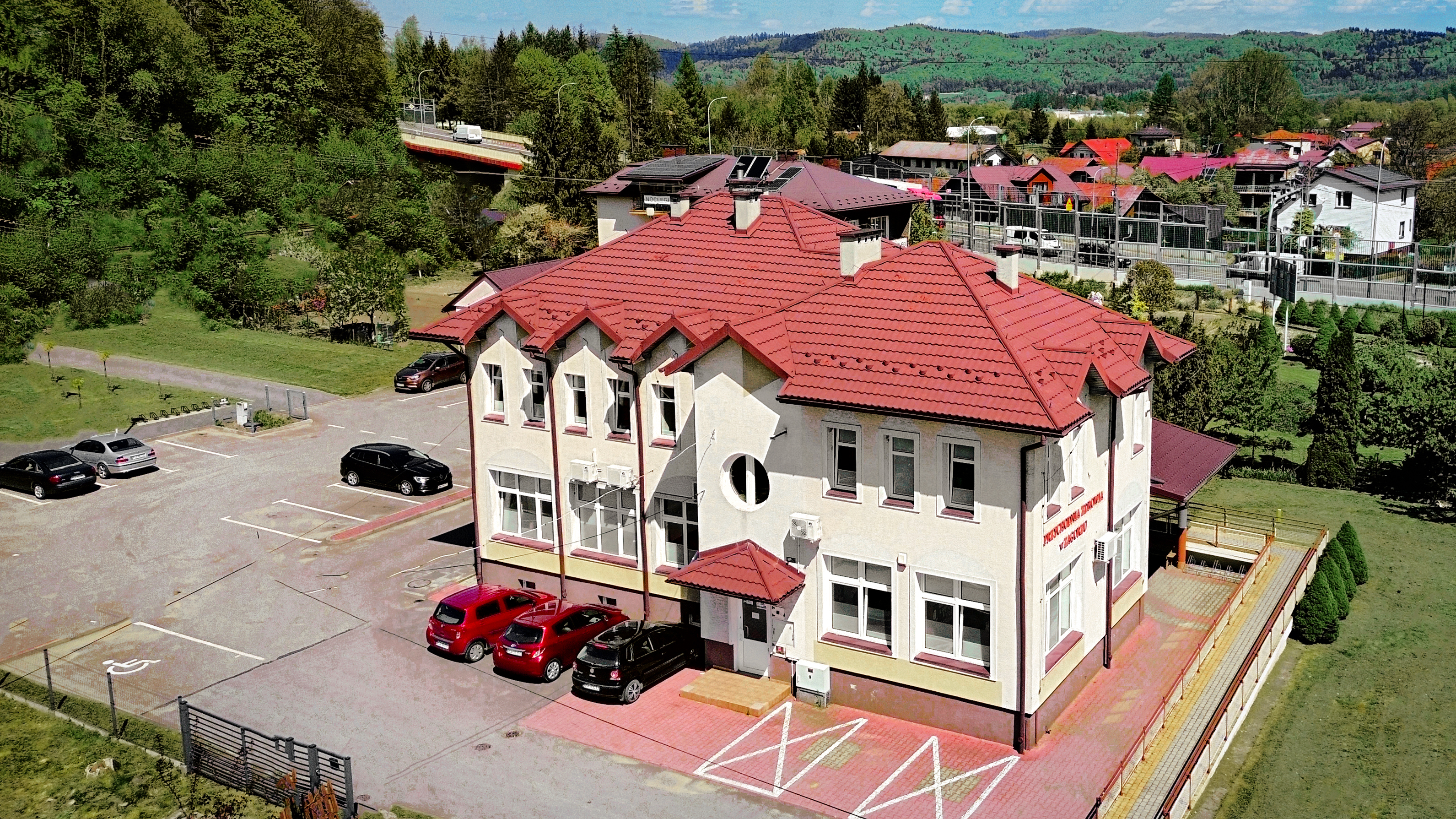 Zdjęcie budynku z czerwoną dachówką i parkingu, z zielonymi wzgórzami w tle.